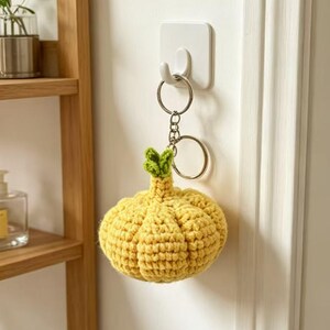 May include: A yellow crocheted pumpkin keychain featuring a small green stem at the top. The pumpkin is attached to a silver-toned metal split ring and a short chain, hanging from a white adhesive wall hook mounted on a plain white wall surface.