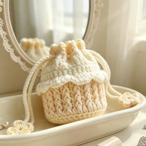 May include: A cream-colored crochet drawstring pouch featuring a textured cable knit pattern on the body, a ruffled edge near the top, and two long braided cords finished with small, six-petaled flower-shaped accents at the ends.