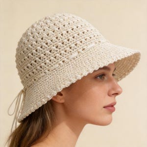 May include: A cream-colored crochet bucket hat with a small brim and a tie closure. The hat has an open weave design, allowing for breathability. The hat is a stylish accessory for sun protection.