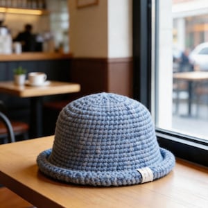 Może przedstawiać: Jasnoniebieski szydełkowy kapelusz typu bucket hat z zaokrągloną główką i krótkim, podwiniętym rondem. Kapelusz ma teksturowany, jednolity wzór ściegu na całej powierzchni oraz małą, prostokątną białą metkę z tkaniny przymocowaną do ronda.
