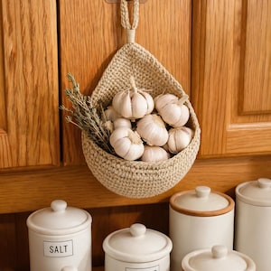 May include: A beige crocheted hanging basket filled with several white garlic bulbs and a sprig of green rosemary. The basket features a single loop handle suspended from a clear adhesive hook. Below are several white ceramic canisters with wooden lids.