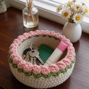 Puede incluir: Una cesta redonda tejida a mano con una base de color crema, una sección central verde con patrón de hojas y un borde rosa inspirado en flores. Dentro de la cesta hay un juego de llaves de metal, un pequeño estuche rectangular verde y un tubo rosa y blanco.