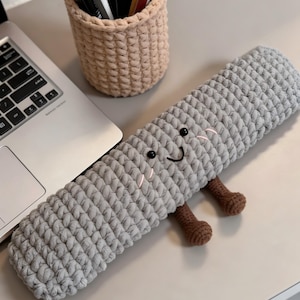 Könnte beinhalten: Ein graues, gehäkeltes Federmäppchen mit einem lächelnden Gesicht und kleinen braunen Beinen. Das rechteckige Federmäppchen liegt auf einer hellen Oberfläche neben einem Laptop und einem beigefarbenen, gehäkelten Stifthalter.