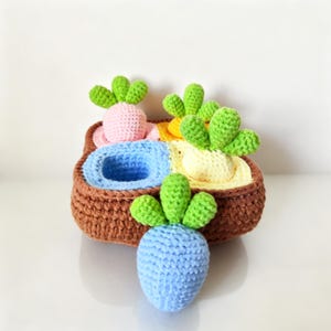 May include: A handmade crochet basket with four compartments, each holding a different coloured crochet vegetable. The vegetables include pink, yellow, and blue carrots, and a green-topped white carrot. The basket is brown.