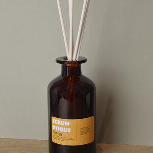 May include: A brown glass diffuser bottle with white reeds. The bottle has a yellow label with the text "Scrumptious Energy". The diffuser is designed to release fragrance into the air.