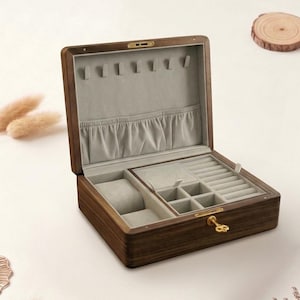 Może przedstawiać: Otwarta drewniana szkatułka na biżuterię z jasnoszarym wnętrzem. Pudełko jest prostokątne z wieloma przegródkami na pierścionki, zegarki i inną biżuterię. Pudełko wykonane jest z ciemnobrązowego drewna i ma złote zapięcie.