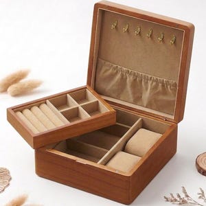 Puede incluir: Una caja de joyería de madera con tapa con bisagras, abierta para revelar múltiples compartimentos forrados con tela beige. La caja tiene una bandeja extraíble para anillos y objetos pequeños. Ganchos dorados montados dentro de la tapa.