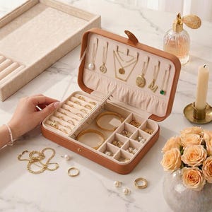May include: Open brown jewelry box with various gold necklaces, rings, bracelets, and earrings. The box is filled with jewelry, including a pearl necklace, gold rings, and a gold bracelet. A tray and a perfume bottle are also visible.