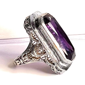 May include: A silver-toned ring featuring an elongated, rectangular, faceted purple gemstone set in a decorative, openwork metal frame with intricate filigree detailing along the sides and a polished, metallic finish on the band structure.