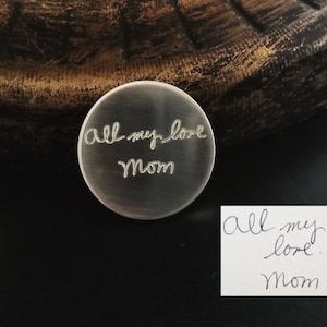 Op de afbeelding: Een kleine, ronde, zilverkleurige metalen herinnering met de woorden "all my love mom" gegraveerd. Een handgeschreven notitie met dezelfde tekst ligt ernaast. De achtergrond is donker.