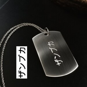 May include: A silver dog tag pendant with a matching chain. The tag has Japanese characters engraved on it. The pendant is rectangular with rounded corners and hangs from a delicate chain. The background is dark.