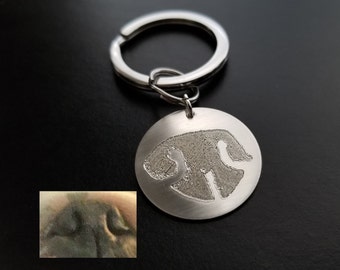 May include: A silver keychain with a round tag featuring a black and white silhouette of a dog. The tag is attached to a silver key ring.