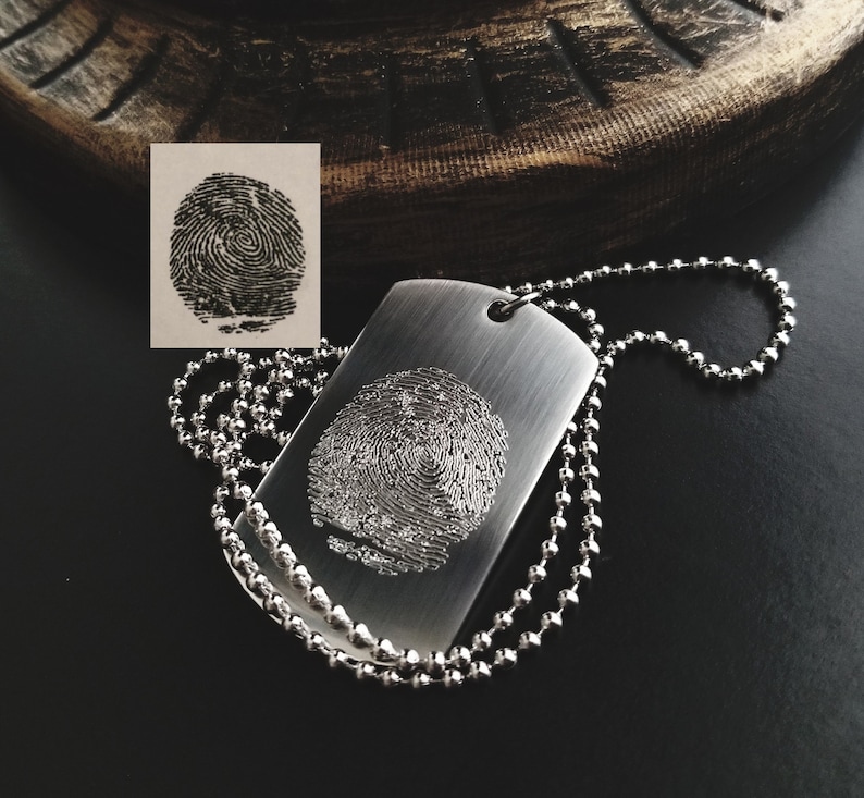 May include: A silver dog tag necklace with a fingerprint design engraved on the front. The necklace is made of a silver chain.