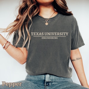 Può includere: Una maglietta grigio scuro con la scritta "TEXAS UNIVERSITY ENGINEERING" in beige chiaro. La maglietta è a girocollo e sembra essere realizzata in un materiale morbido e confortevole. La parola "Pepper" è in basso a sinistra.