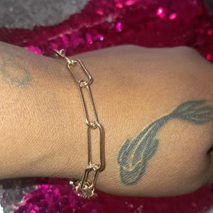 May include: A gold-colored link bracelet. The bracelet is made of elongated rectangular links. The bracelet is on a wrist with a tattoo. The background is a pink sequined surface.