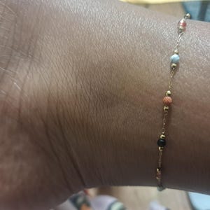 May include: A delicate gold anklet with small, colourful beads. The anklet's chain is thin and the beads are in shades of red, orange, blue, and black. The anklet is worn on the ankle.