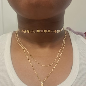 Puede incluir: Conjunto de collares dorados superpuestos. El superior es un choker con pequeños discos redondos. El segundo es una cadena delicada, y el tercero presenta un colgante de cruz. Llevado sobre una camiseta sin mangas blanca.
