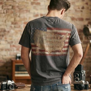 May include: Dark grey t-shirt with a distressed American flag design and a map of the United States. The short-sleeved shirt has a vintage look. The person is wearing blue jeans.