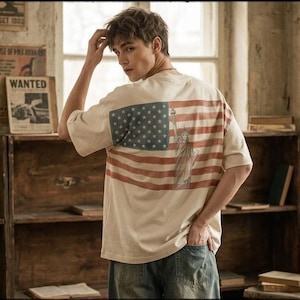 May include: Cream-coloured t-shirt with the American flag design on the back. The flag has red and white stripes and a blue field with white stars. The Statue of Liberty is printed on the flag. The shirt is worn with blue jeans.