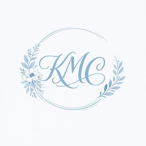 May include: A circular logo design featuring the letters KMC in a blue cursive script, centered within a delicate wreath of blue leaves and a single flower on the left side, all set against a plain white background for a clean visual presentation.