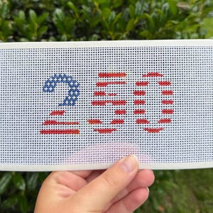 May include: A rectangular white needlepoint canvas featuring the number 250 stitched in a patriotic style. The number 2 is filled with blue stars, while the numbers 5 and 0 are composed of horizontal red stripes. The canvas has white taped edges.