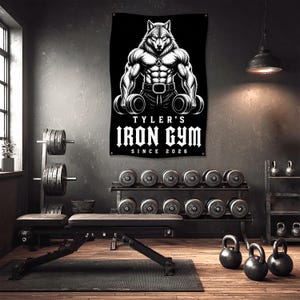 May include: Black banner with a muscular wolf holding dumbbells, the text "TYLER'S IRON GYM SINCE 2026" in white. The gym setting includes weights, a bench, and kettlebells, creating a motivational fitness environment.
