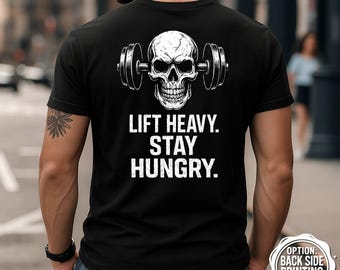 Puede incluir: Camiseta negra con un diseño de calavera blanca sosteniendo una barra y el texto "LIFT HEAVY. STAY HUNGRY." impreso debajo. La parte posterior de la camiseta también tiene un pequeño gráfico circular que indica que la impresión en la parte posterior está disponible.