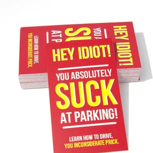 May include: Red cards with bold yellow text that reads "HEY IDIOT! YOU ABSOLUTELY SUCK AT PARKING!" and "LEARN HOW TO DRIVE, YOU INCONSIDERATE PRICK." Stack of cards on a white background.