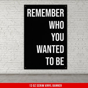 May include: Black vinyl banner with white text that reads "Remember who you wanted to be". The banner is hanging on a white brick wall. 13 oz scrim vinyl banner.