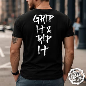 May include: Black t-shirt with the white text "GRIP IT & RIP IT" vertically aligned on the back. The shirt is a crew neck style. A small tattoo is visible on the left arm. The image is taken outdoors.