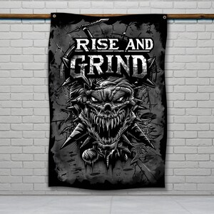 Puede incluir: Bandera negra y gris con las palabras "RISE AND GRIND". La bandera presenta una ilustración detallada de una calavera demoníaca con dientes afilados y púas, creando una estética oscura e intensa. La bandera está colgada.