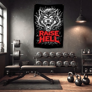 May include: Black wall flag featuring a detailed white illustration of a snarling wolf's head. Bold red text below reads "RAISE HELL". The flag hangs in a gym setting with weights and exercise equipment.
