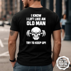 Könnte beinhalten: Schwarzes T-Shirt mit weißem Text und einem Totenkopf-Motiv, das eine Langhantel hält. Der Text lautet: "I KNOW I LIFT LIKE AN OLD MAN TRY TO KEEP UP!". Ein kleines Grafik sagt "OPTION: BACK SIDE PRINTING AVAILABLE."