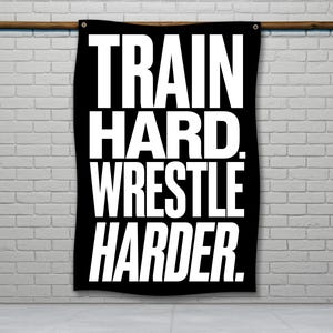 May include: Black banner with the words "TRAIN HARD. WRESTLE HARDER." in bold white letters. The banner is hanging on a wooden rod against a white brick wall. The banner is rectangular and has grommets in the top corners.