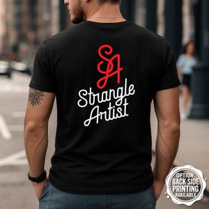 May include: Black t-shirt with the text "SA Strangle Artist" in white script and a red stylized "SA" logo. The shirt is a crew neck style and is being worn by a person.