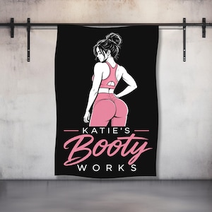 May include: Black fitness towel featuring a pink illustration of a woman in workout attire. The towel has the text "KATIE'S Booty WORKS" in pink script. The woman is wearing a pink sports bra and leggings.