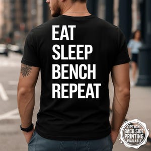 Pode incluir: Camiseta preta com o texto branco "EAT SLEEP BENCH REPEAT" impresso nas costas. A camiseta é de gola redonda. A imagem também inclui um pequeno gráfico circular que diz "OPTION: BACK SIDE PRINTING AVAILABLE".