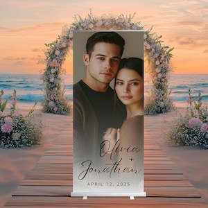 May include: A wedding backdrop with a photo of a couple in front of a beach sunset. The backdrop is white with black text that reads "Olivia + Jonathan" and "April 12, 2025".