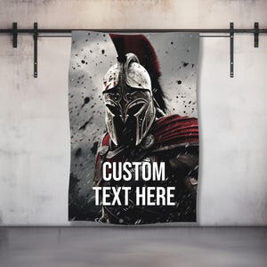 Peut inclure: Une tenture murale représentant un guerrier avec un casque et une cape rouge, avec le texte "CUSTOM TEXT HERE" en blanc. L'œuvre d'art a une palette de couleurs grise et noire, avec l'accent mis sur le visage et l'armure du guerrier.