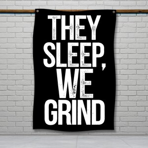 Puede incluir: Bandera negra con el texto blanco "THEY SLEEP, WE GRIND". La bandera está colgada de una barra de madera contra una pared de ladrillos blancos. El texto está en una fuente en negrita, sans-serif.