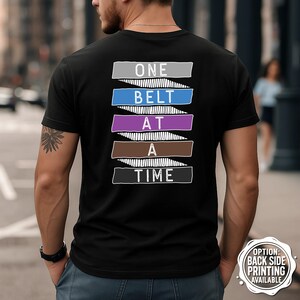 May include: Black t-shirt featuring the text "ONE BELT AT A TIME" in white, with coloured bars representing martial arts belt colours. The shirt is being worn by a person.