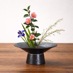 May include: A black ceramic ikebana vase displays a floral arrangement. The arrangement features pink camellias, blue irises, and white flowering branches. The vase has a wide, shallow bowl and a textured pedestal base, set on a wooden surface.