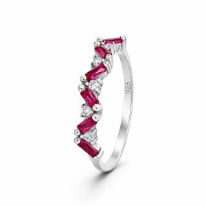 May include: A silver ring featuring a chevron-shaped band set with alternating baguette-cut red stones and round clear stones. The inner band is clearly stamped with the number 925, indicating sterling silver composition for this jewellery piece.