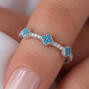 May include: A silver-toned ring featuring a series of small, four-petaled flower motifs. Each flower is composed of four tiny, round, bright turquoise-colored stones. The band is further accented with small, clear, sparkling white stones.