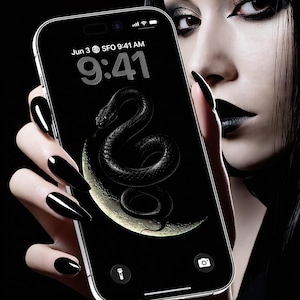 May include: A black smartphone displays a dark wallpaper featuring a coiled snake on a crescent moon. The screen shows the time as 9:41 AM on June 3rd. The phone is held by a person with black nails and dark makeup.