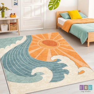 May include: A rectangular rug with a stylized design featuring a large orange sun and blue waves. The rug has a cream-colored background and is placed on a light-colored floor. The rug is in a room with a bed and a side table.