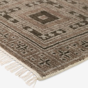 May include: Close-up of a brown and beige geometric patterned rug. The rug features a repeating diamond design with darker brown accents. The edge has a cream-colored border with fringe.