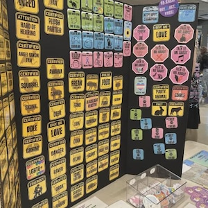 Puede incluir: Una exhibición de pegatinas coloridas con varios diseños y texto, incluyendo ilustraciones de animales y frases como "Certificado", "Amor" y "Animal de fiesta". Las pegatinas están dispuestas en un tablero de exhibición negro.