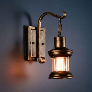 May include: Antique-style wall sconce with a bronze finish. The fixture features a rectangular wall mount, a curved arm, and a lantern-style light with clear glass panels. The light is illuminated, casting a warm glow.