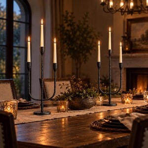 May include: Two black metal candelabras with a textured finish, each holding three lit white taper candles. The candelabras are placed on a long wooden dining table alongside small glass votive candle holders and a central bowl of greenery.